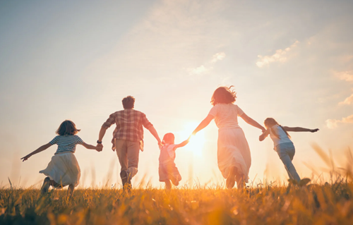 image shows the backs of two adults and three children holding hands, running in a field as the sun sets